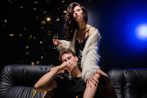 Donna sexy in abito e giacca bianca seduta sul divano dietro l'uomo che beve champagne, mentre i coriandoli cadono, con retroilluminazione sul nero in discoteca — Foto stock