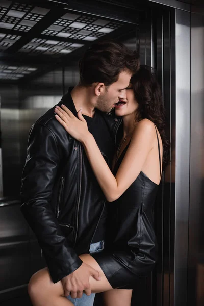 Man in leather jacket kissing and holding leg of seductive woman in black dress in elevator — Stock Photo