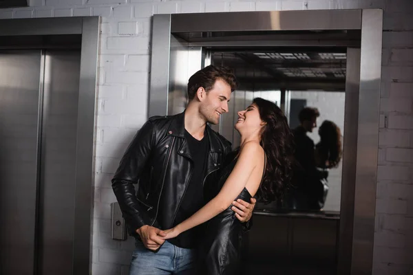 Улыбающийся мужчина в черной куртке обнимает и держит за руку веселую кудрявую женщину в кожаном платье у входа в лифт — стоковое фото