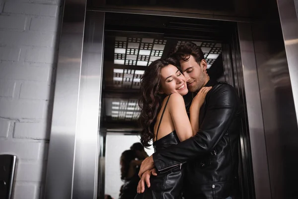 Hombre sonriente con los ojos cerrados abrazando mujer morena feliz en la entrada del ascensor - foto de stock