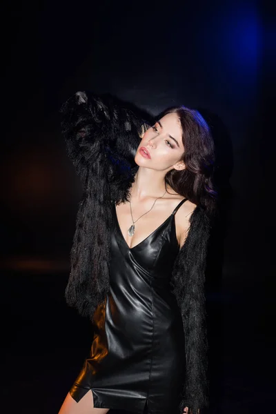 Seductive brunette woman in leather dress and jacket posing with hand behind head, while standing and looking away on black — Stock Photo