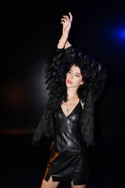 Sexy brunette woman with hands in air, wearing leather dress and jacket, looking away, while standing on black — Stock Photo