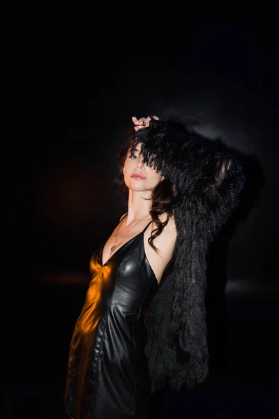 Sexy curly woman posing with hand near head, looking at camera, while wearing jacket and leather dress on black — Stock Photo