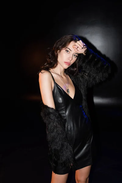 Curly brunette woman in jacket and leather dress posing with hand near head, while looking at camera on black — Stock Photo