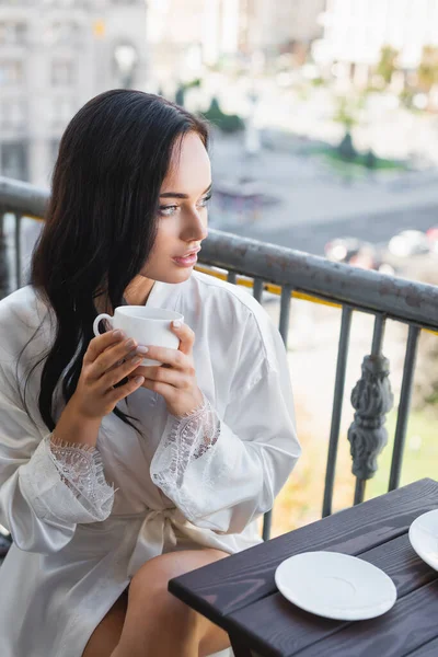 Красива брюнетка в білому халаті п'є каву на балконі — стокове фото