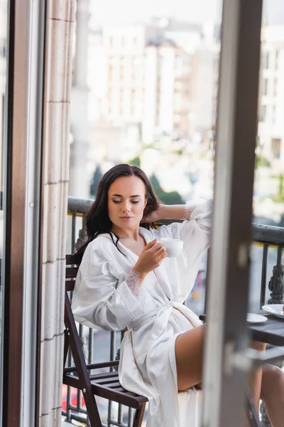 Beautiful brunette woman in white robe drinking coffee on balcony — Stock Photo