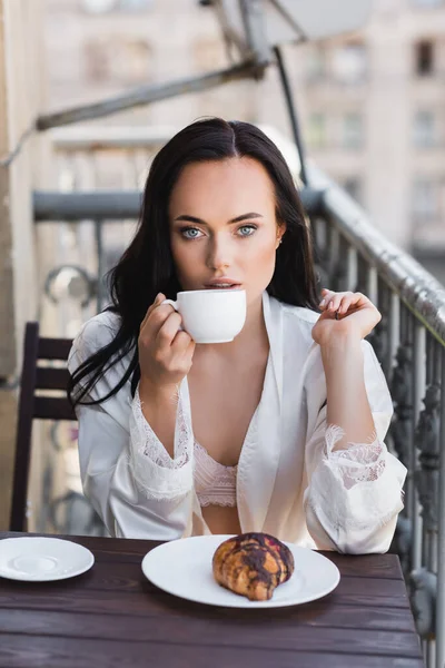 Beautiful brunette woman in white robe drinking coffee on balcony — Stock Photo