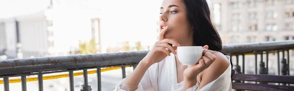 Femme brune en robe blanche buvant du café et tenant les doigts près de la bouche, bannière horizontale — Photo de stock