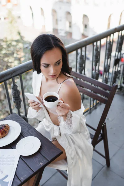 Hermosa mujer morena en bata blanca sosteniendo teléfono inteligente y beber café en el balcón - foto de stock