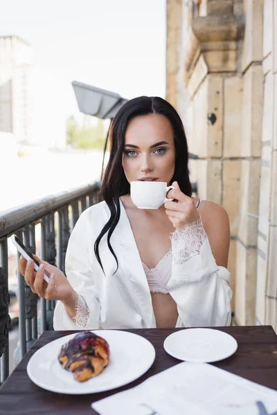 Belle femme brune en robe blanche tenant smartphone et prenant le petit déjeuner sur le balcon — Photo de stock