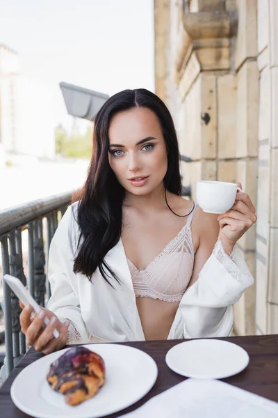 Belle femme brune en robe blanche tenant smartphone et prenant le petit déjeuner sur le balcon — Photo de stock