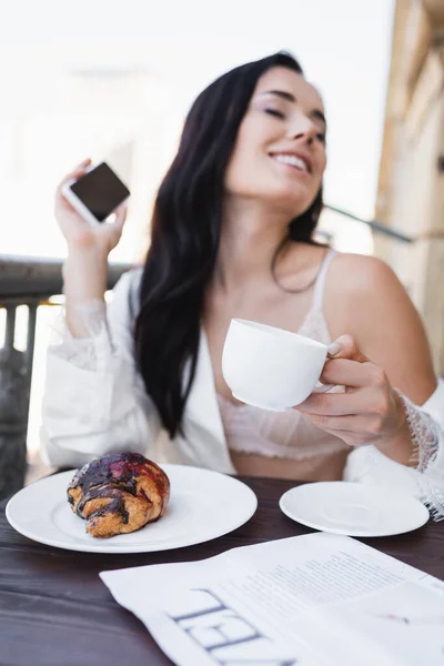 Beautiful brunette woman in white robe holding smartphone and having breakfast on balcony — Stock Photo