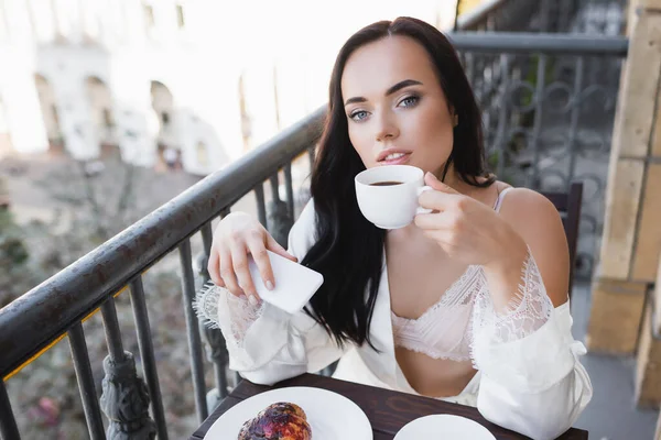 Bela mulher morena em roupão branco bebendo café na varanda e segurando smartphone — Fotografia de Stock