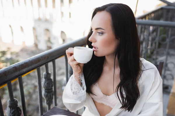 Hermosa mujer morena en túnica blanca bebiendo café y mirando hacia otro lado - foto de stock