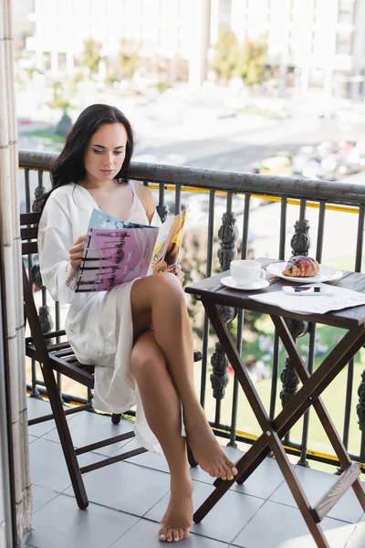 Брюнетка в белом халате сидит на балконе и читает журнал — стоковое фото