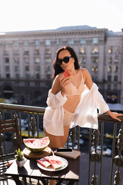Morena mujer en ropa interior beige y camisa blanca comiendo sandía en balcón - foto de stock