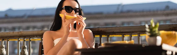 Sexy femme brune en lunettes de soleil et recouvert de feuille blanche manger melon sur le balcon, bannière horizontale — Photo de stock