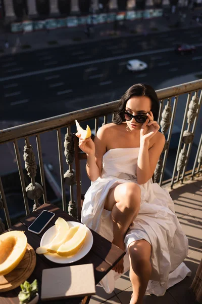 Sexy brunette woman raising sunglasses and eating melon on balcony — Stock Photo