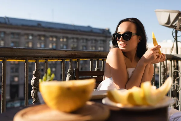 Sexy bruna donna in occhiali da sole e coperto di lenzuolo bianco mangiare melone sul balcone — Foto stock