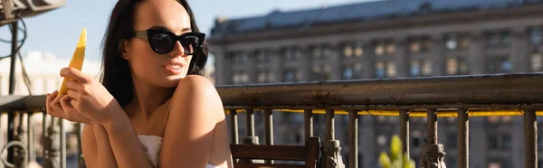 Сексуальна брюнетка в сонцезахисних окулярах і покрита білим листом, їсть диню на балконі, горизонтальний банер — стокове фото