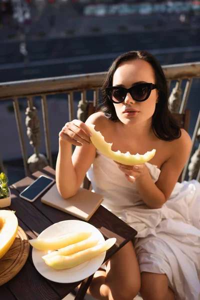 Сексуальная брюнетка в солнечных очках и покрытая белым полотном, поедающая дыню на балконе — стоковое фото