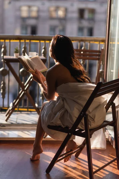 Back view of attractive woman covered in white sheet reading book and sitting on balcony — Stock Photo