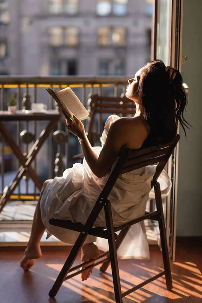 Attractive woman covered in white sheet reading book and sitting on balcony — Stock Photo
