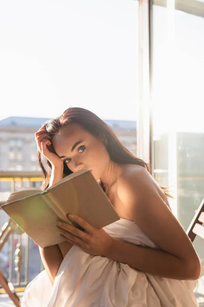 Attraktive Frau mit weißem Laken bedeckt, liest Buch und sitzt auf Balkon — Stockfoto