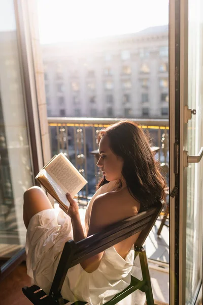 Attractive woman covered in white sheet reading book and sitting on balcony — Stock Photo