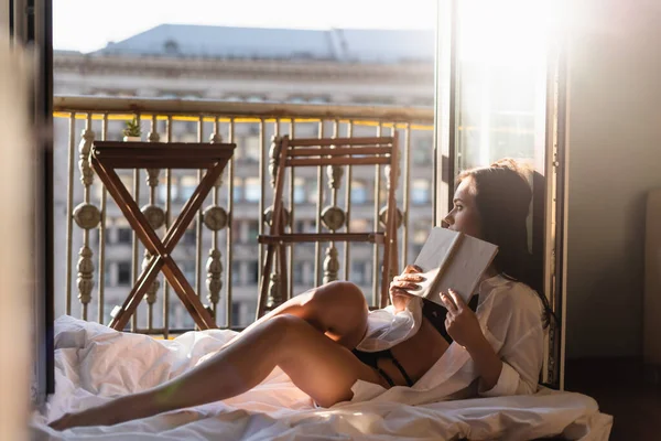 Mulher atraente na camisa branca segurando livro e sentado em cobertor — Fotografia de Stock