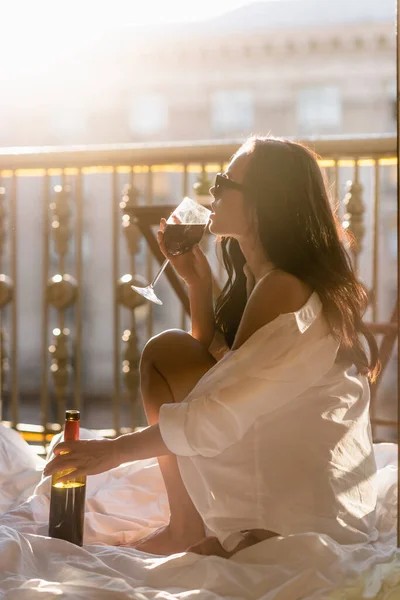 Brunette woman in sunglasses and white shirt drinking wine on balcony — Stock Photo