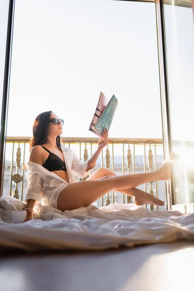 Schöne brünette Frau mit Sonnenbrille und schwarzer Unterwäsche, die Zeitschrift liest und eine Tasse Tee hält — Stockfoto