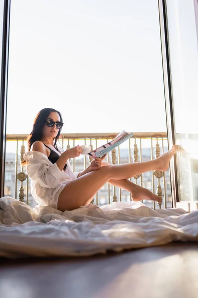 Hermosa morena mujer en gafas de sol y ropa interior negro lectura revista y beber té - foto de stock