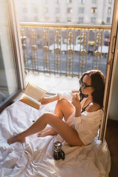Bella donna bruna in occhiali da sole leggere libro e bere tè seduto sulla coperta — Foto stock
