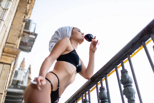 Sexy young woman in black underwear with towel on head smoking cigarette and drinking red wine on balcony — Stock Photo