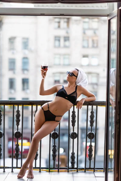 Sexy young woman in black underwear with towel on head drinking red wine on balcony — Stock Photo