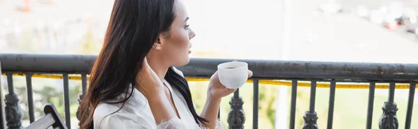 Брюнетка в білому халаті п'є чай і сидить на балконі, горизонтальний банер — стокове фото