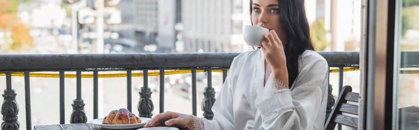 Morena mujer en túnica blanca bebiendo té y sentado en el balcón, pancarta horizontal - foto de stock