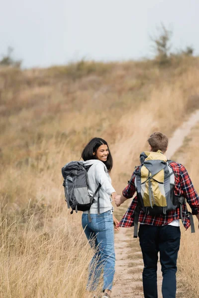 Femme afro-américaine souriante avec sac à dos tenant la main du petit ami pendant la randonnée sur le chemin — Photo de stock