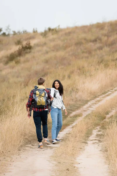 Smiling african american woman looking at boyfriend with backpack while walking on path during trip — Stock Photo