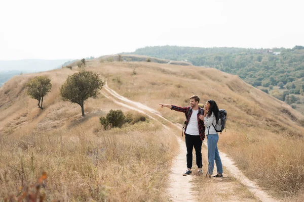 Smiling man with backpack pointing with finger near excited african american girlfriend on path during trip — Stock Photo