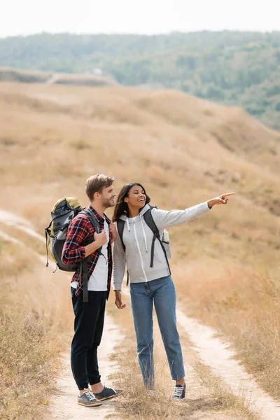 Donna afroamericana che punta con il dito vicino al fidanzato sorridente con lo zaino sul sentiero all'aperto — Foto stock