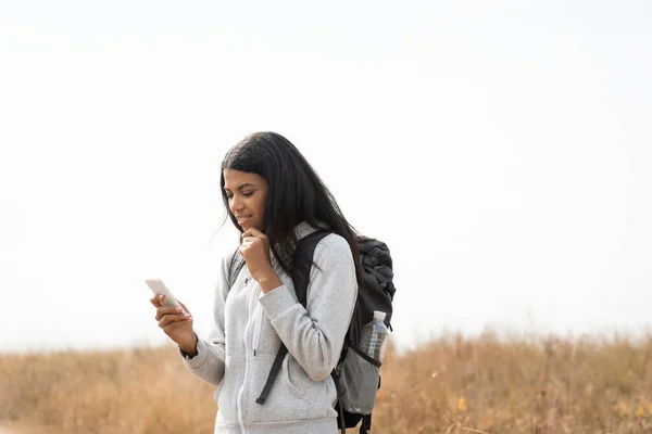 Pensiva donna afroamericana con zaino utilizzando smartphone all'aperto — Foto stock