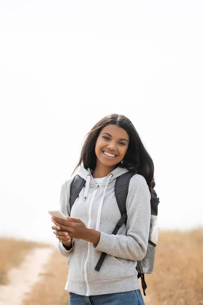 Sorrindo afro-americano turista segurando smartphone enquanto olha para a câmera ao ar livre — Fotografia de Stock