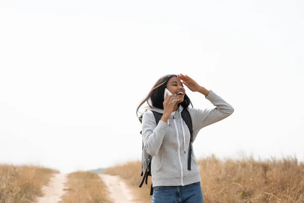 Viajero afroamericano positivo hablando en un teléfono inteligente cerca del camino sobre un fondo borroso - foto de stock