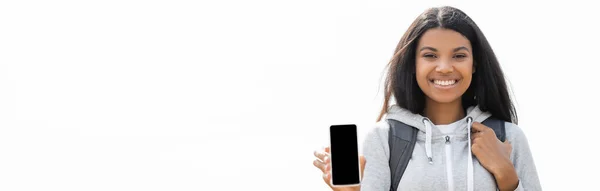 Smiling african american woman with backpack showing smartphone isolated on white, banner — Stock Photo