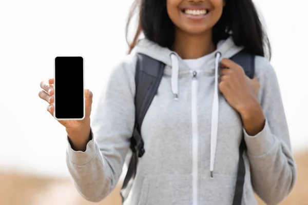 Smartphone with blank screen in hand of african american woman with backpack on blurred background outdoors — Stock Photo