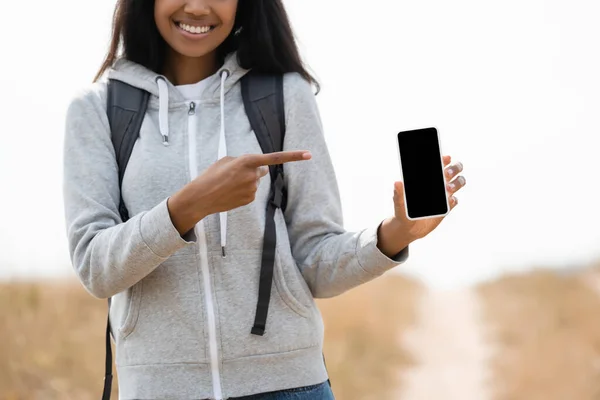 Vista ritagliata di sorridente turista afroamericano che punta con il dito allo smartphone con schermo vuoto all'aperto — Foto stock