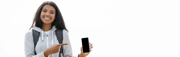 Улыбающийся африканский турист, указывающий пальцем на смартфон с пустым экраном, изолированным на белом, баннер — стоковое фото
