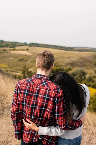 Rückansicht eines multiethnischen Paares, das sich mit Hügeln auf verschwommenem Hintergrund umarmt — Stockfoto